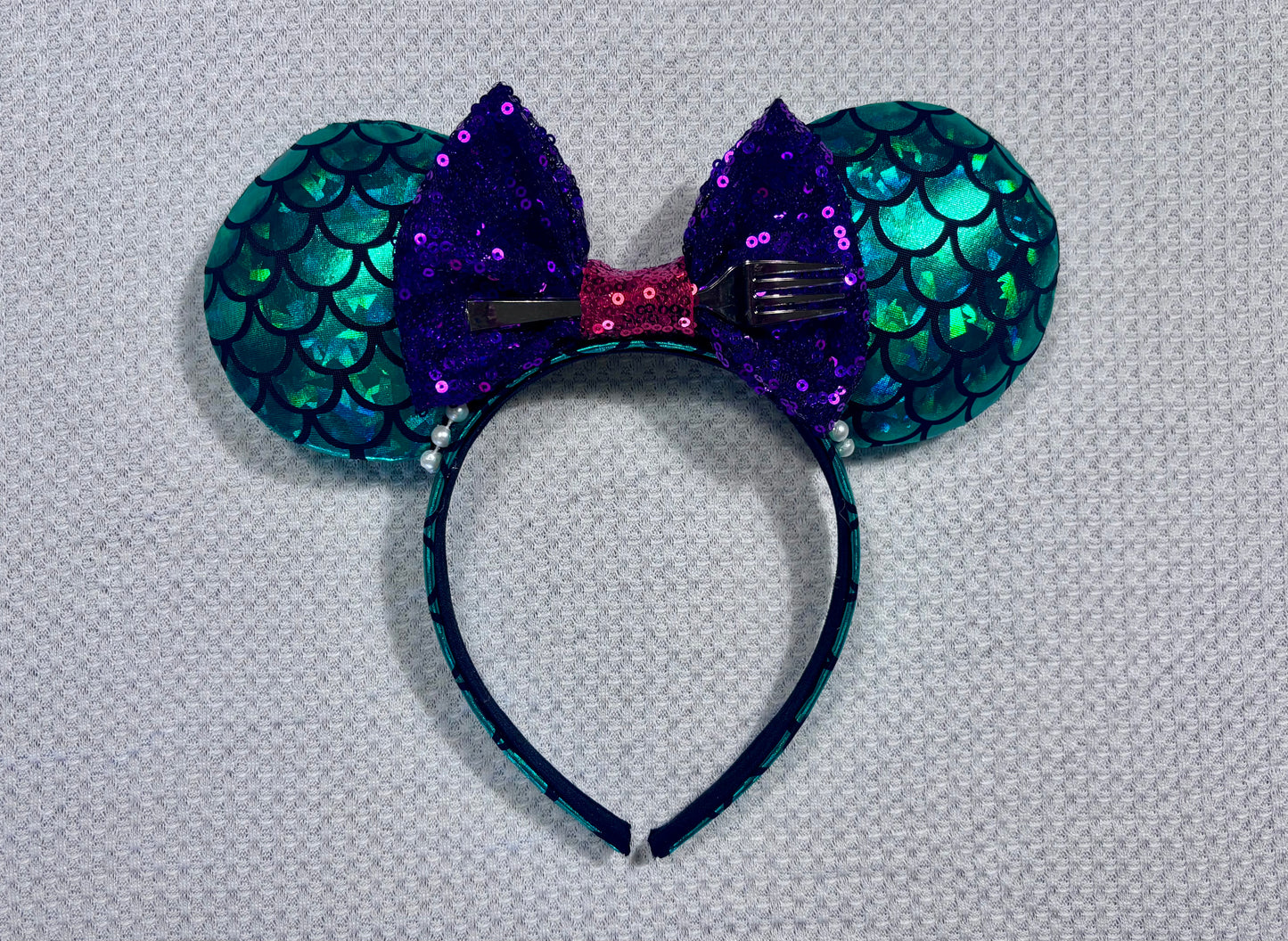 Mermaid-themed headband with teal scales and a purple sequin bow with a red middle and a dinglehopper middle piece. Pearl garland trim along inner ears m on a matching mermaid scales headband