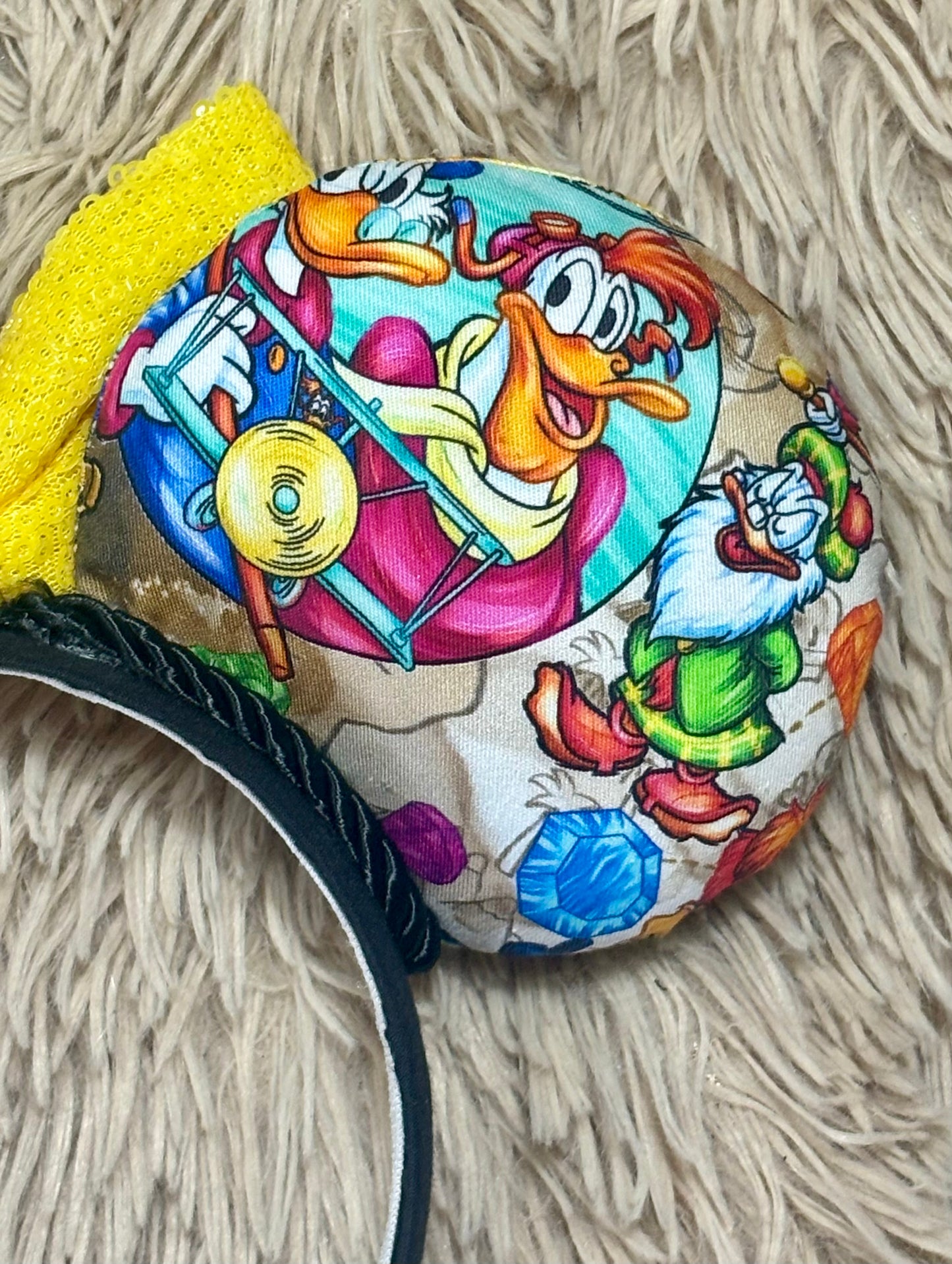 A headband featuring a colorful cartoon print of famous cartoon Duck characters, accented by a large yellow sequined bow in the center. Close up of the back of one of the ears.