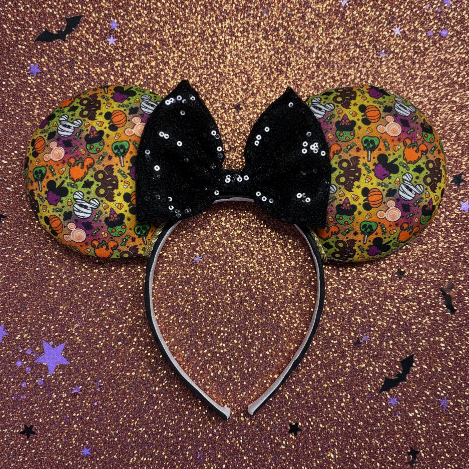 An ear headband featuring a black sequined bow in the center and patterned ears with a colorful Halloween-themed print, displayed on an orange glittery Halloween background.