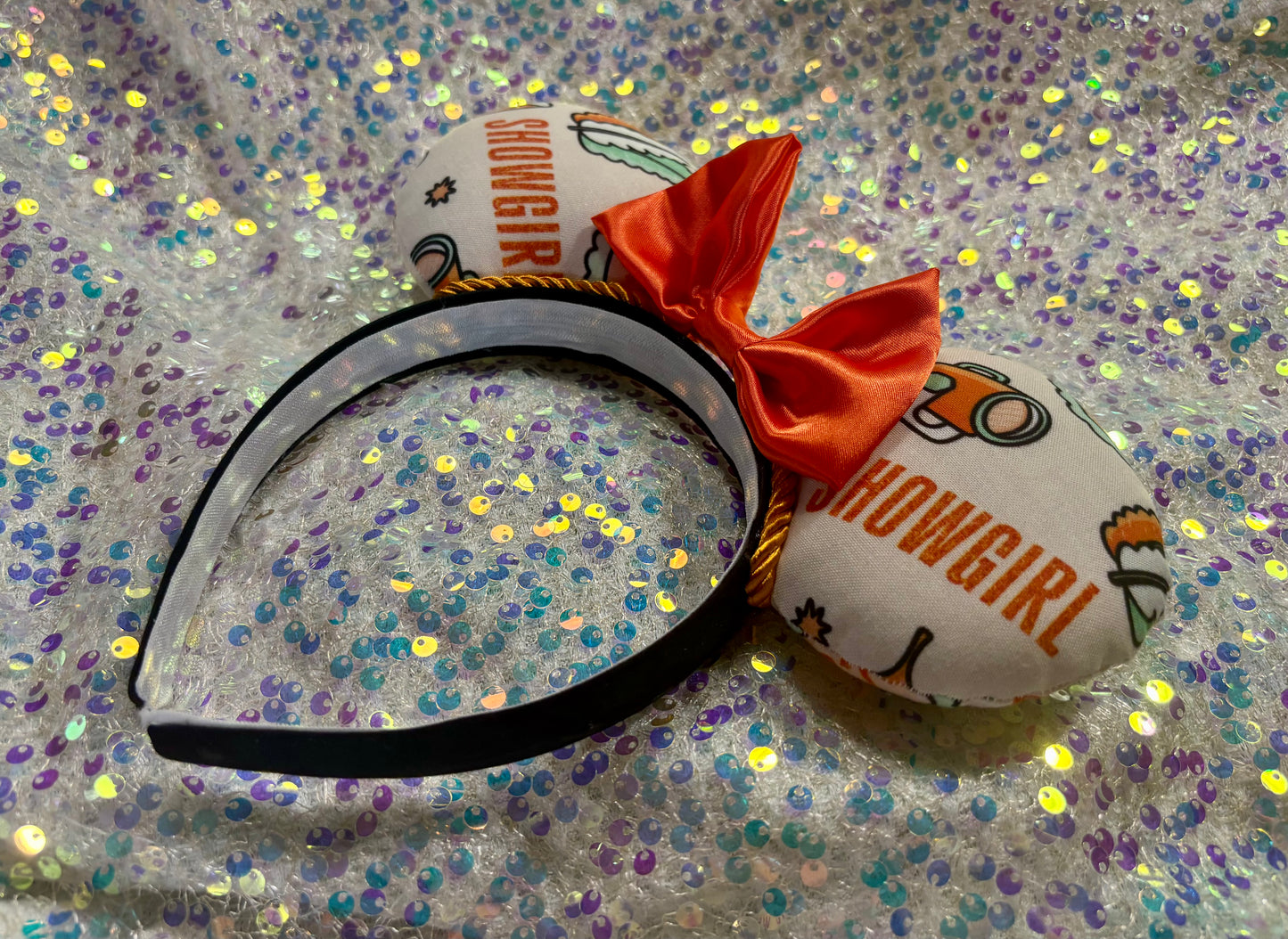 Angled view of headband. Showgirl themed mouse ears with an orange satin bow on a black headband.