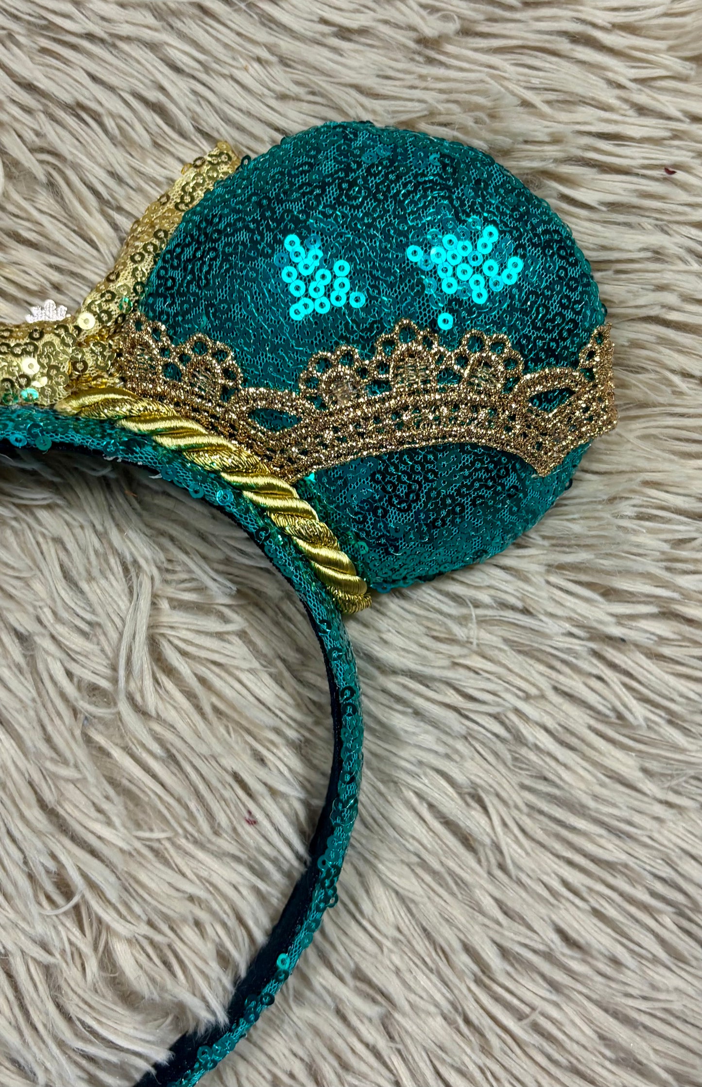 A pair of sequined mouse ears headband featuring teal and gold sequins, a large gold sequin bow with a blue gemstone center, and gold lace trim.