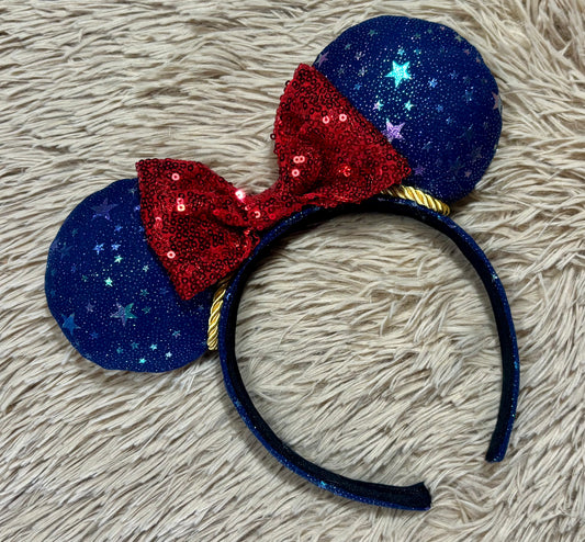An ear headband featuring dark blue glittery ears with silver star patterns, a large red sequined bow, and a dark blue glitter headband, displayed on a beige shag rug.