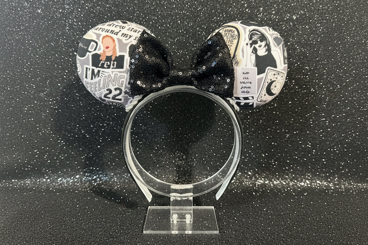 Black and white Swiftie cartoon themed mouse ears with a black sequin bow on a black headband. Shown on a clear stand against a dark background.