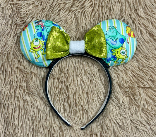 A Monster themed headband with two round ears featuring a light blue and yellow striped pattern with Mike and Sulley prints, accented by a large shiny olive green bow in the center and a white faux-fur trim.