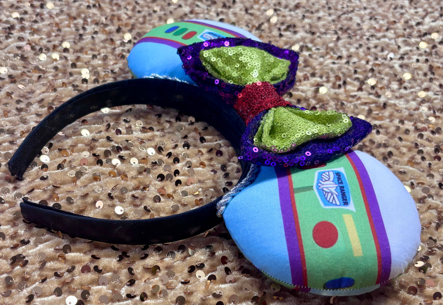 Side view of mouse ears and the headband. Space Ranger Mouse Ear headband with new improved bow. Handmade and ready to ship with Space ranger fabric on both the front and the back of mouse ears. The set has a silver twisted trim along the inner ears and a new sequin bow that is purple, lime green and red in sequin. Ears are on a black poly cotton universal sized headband.