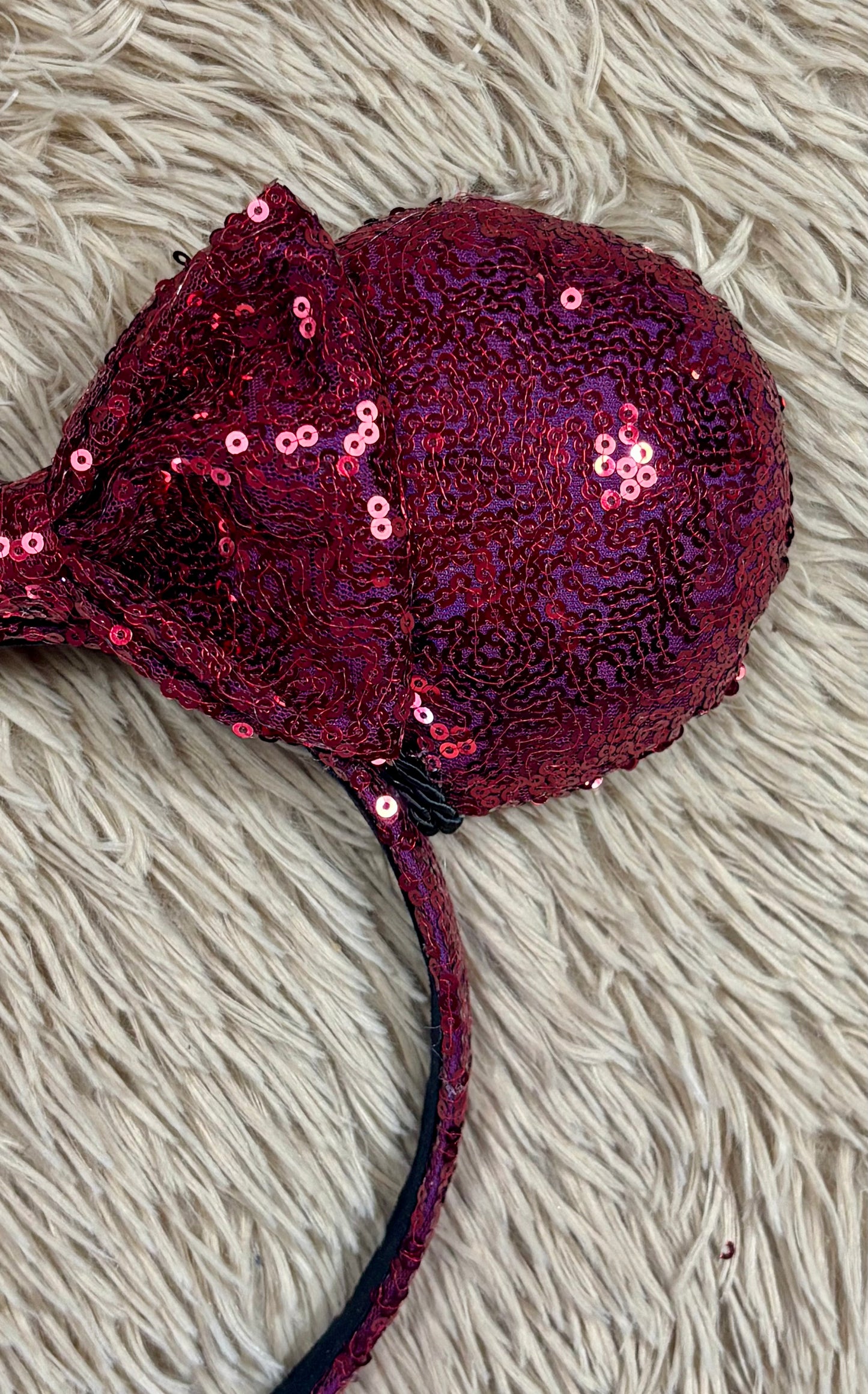 A close-up of a sequined headband featuring a large bow and a round cup-like section covered in dark red and magenta sequins, laid on a beige shag rug.