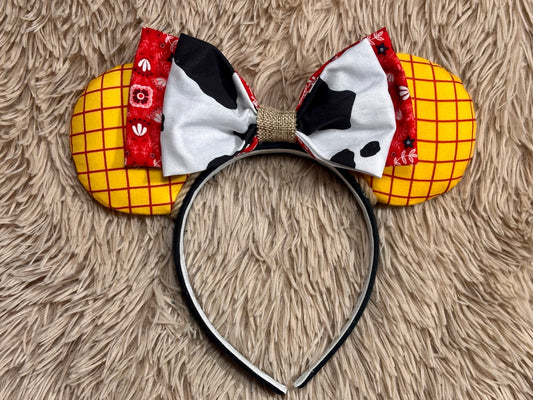 A pair of ears headband accessories featuring yellow grid-patterned ears, a red patterned band, and a bow made of black-and-white cow-print fabric and white fabric with black spots