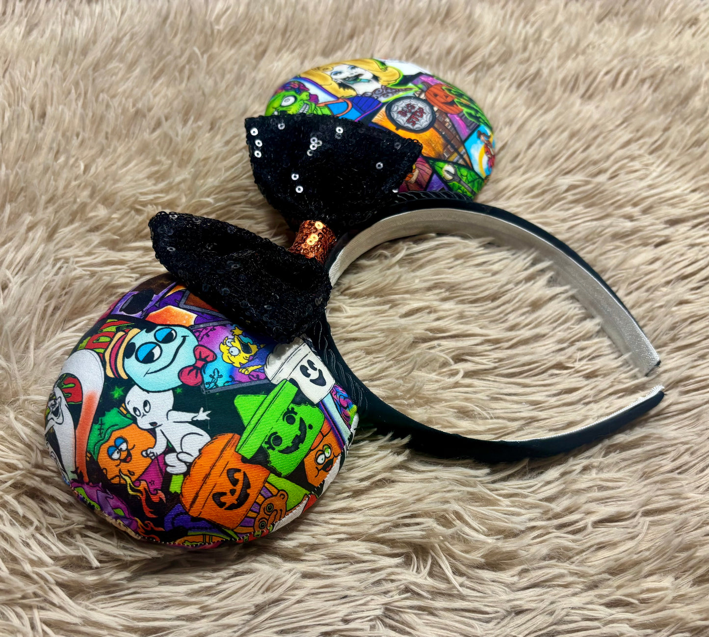 Halloween-themed ear headband featuring a colorful cartoon pattern of ghosts, pumpkins, and bats, with a large black sequined bow, displayed on a beige faux fur background.