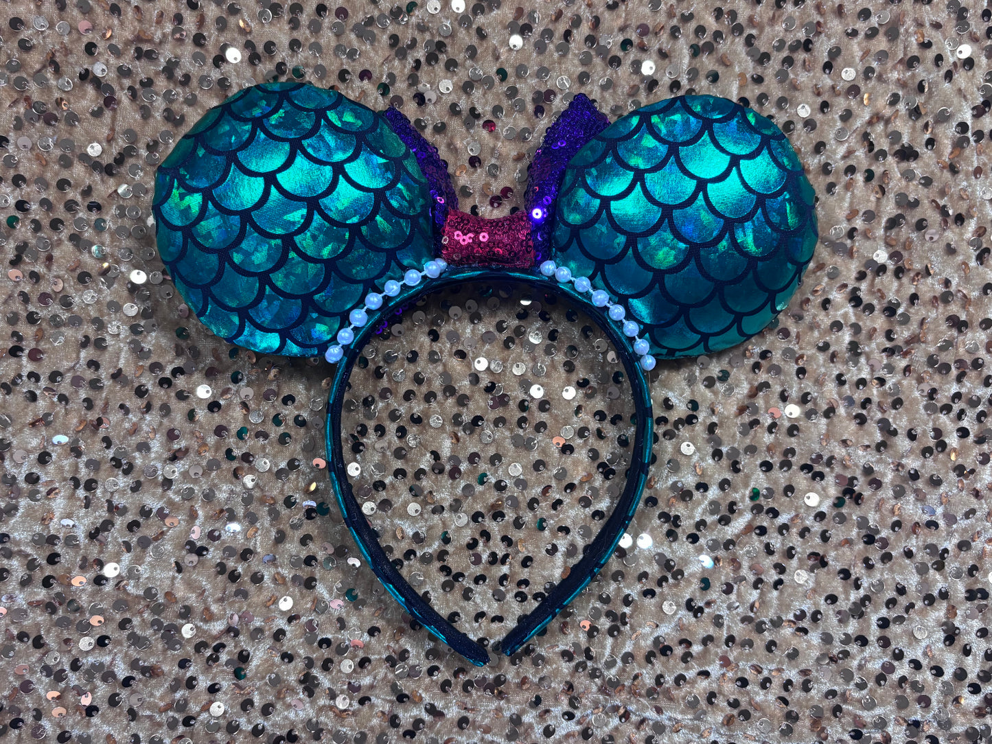 Back of the mouse ears. Mermaid-themed headband with teal scales and a purple sequin bow with a red middle and a dinglehopper middle piece. Pearl garland trim along inner ears m on a matching mermaid scales headband.