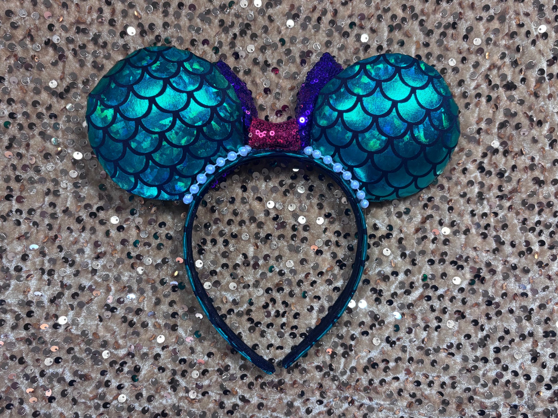 Back of the mouse ears. Mermaid-themed headband with teal scales and a purple sequin bow with a red middle and a dinglehopper middle piece. Pearl garland trim along inner ears m on a matching mermaid scales headband.