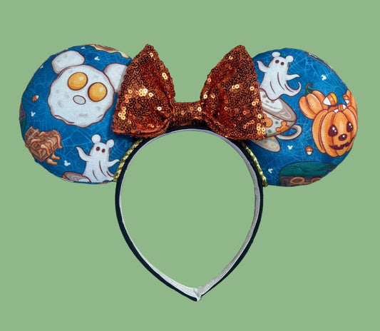 Halloween-themed ears headband featuring blue fabric ears with a breakfast ghost and pumpkin print, a large sequined copper-brown bow, and a black headband.