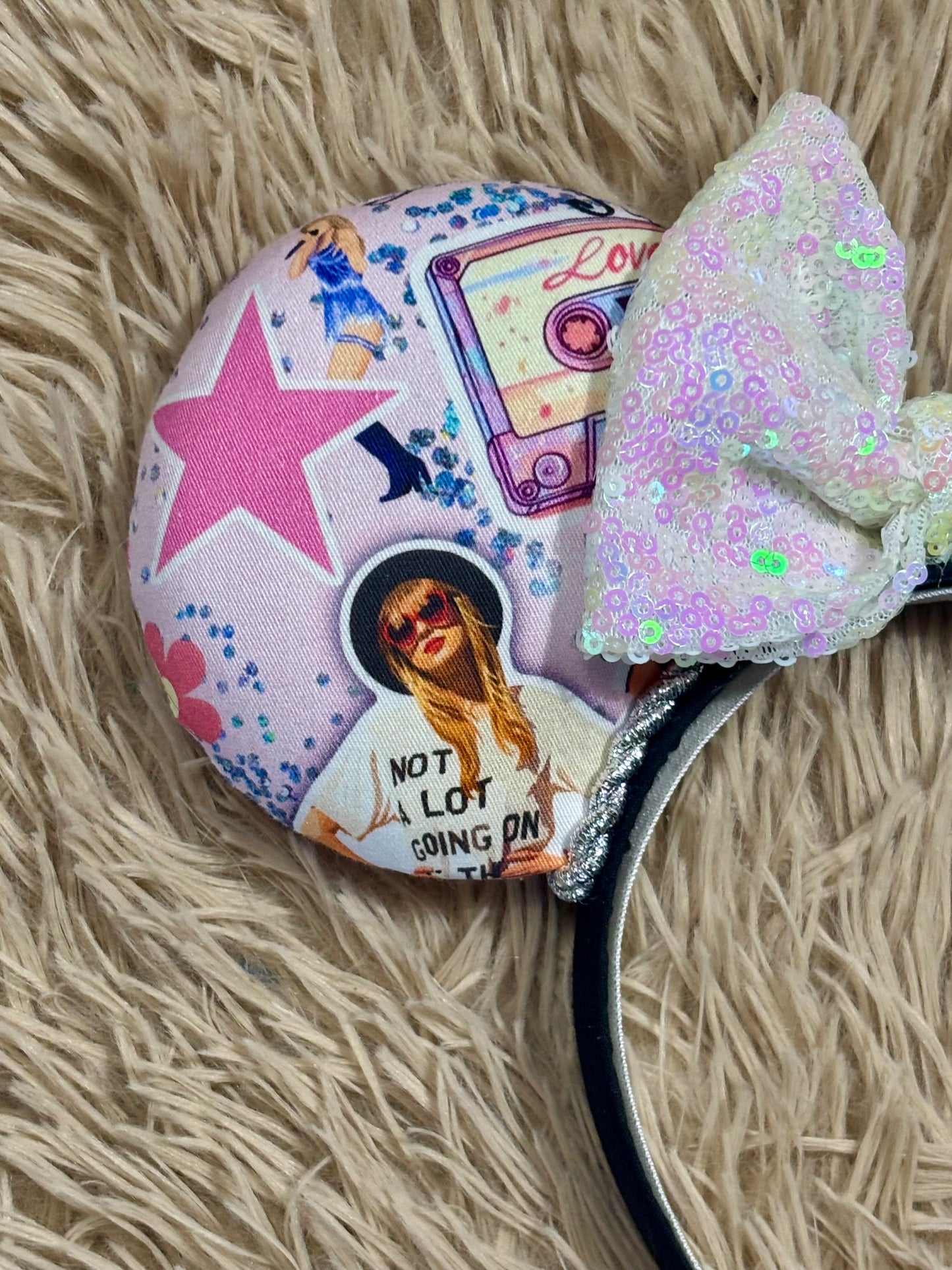 A close-up of a decorative headband featuring a round printed fabric-covered portion with a pink star, cassette tape graphic, collage photo, and text, plus a large iridescent sequined bow on a black band, laid on a beige faux-fur surface.