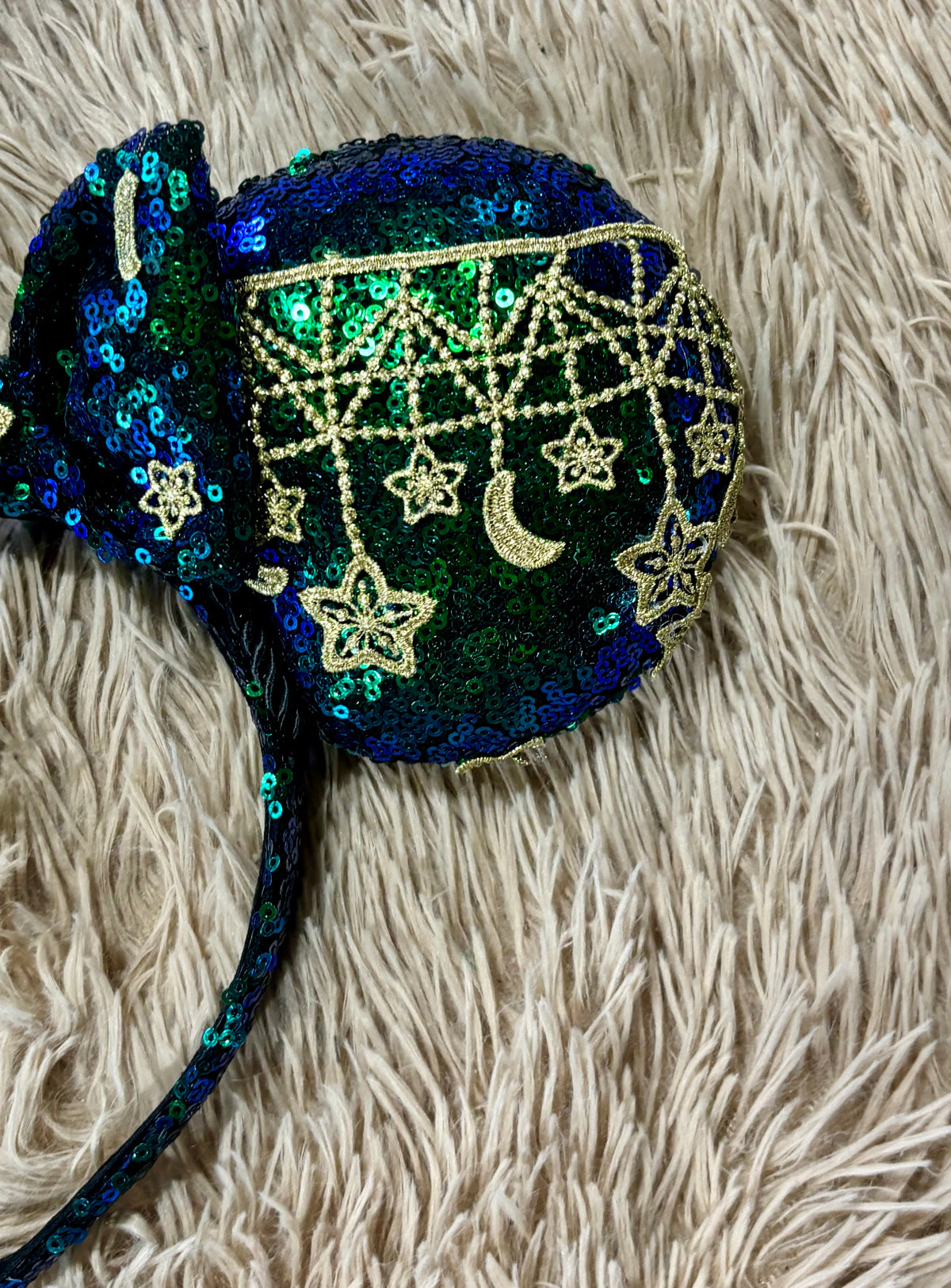 A round sequined headband featuring a dark blue and green iridescent sequin body with gold embroidered stars, a crescent moon, and geometric patterns, resting on a beige faux fur background.