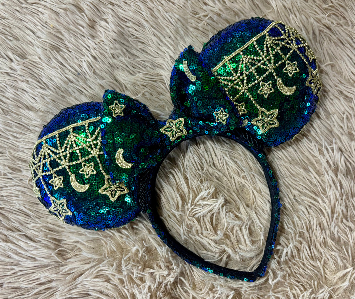 A pair of blue and green iridescent ear headband featuring dark blue and green sequins with gold embroidered stars, moons, and web patterns.