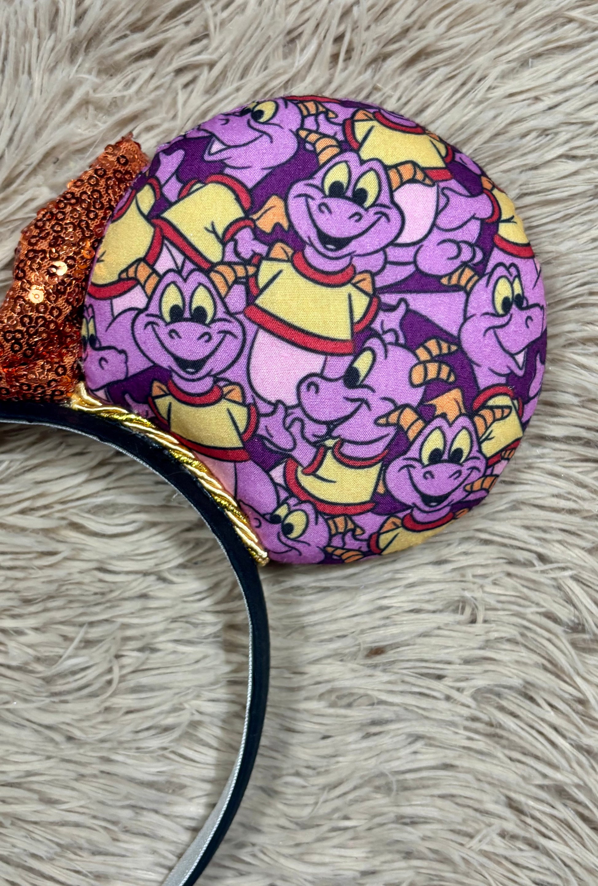 A close-up of a round headband featuring a colorful all-over print of purple cartoon dragons wearing yellow and red outfits, with a glittery orange-brown bow on the left and a black band.