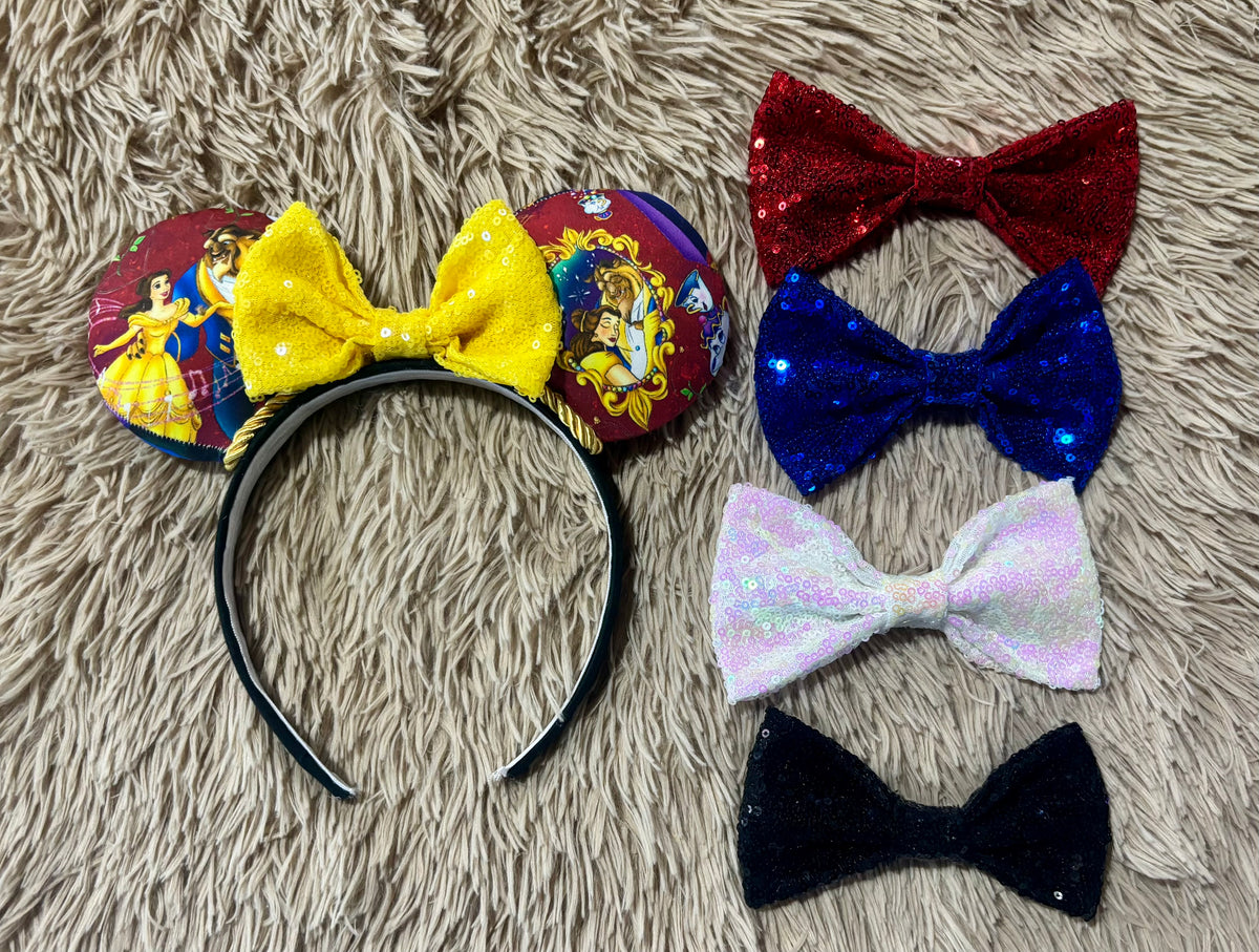 A set of mouse ears laid out on a beige shag rug, including a headband with mouse ears featuring Beauty of the Beast character prints and a large yellow bow. Other bow choices in red, blue, white, and blue.