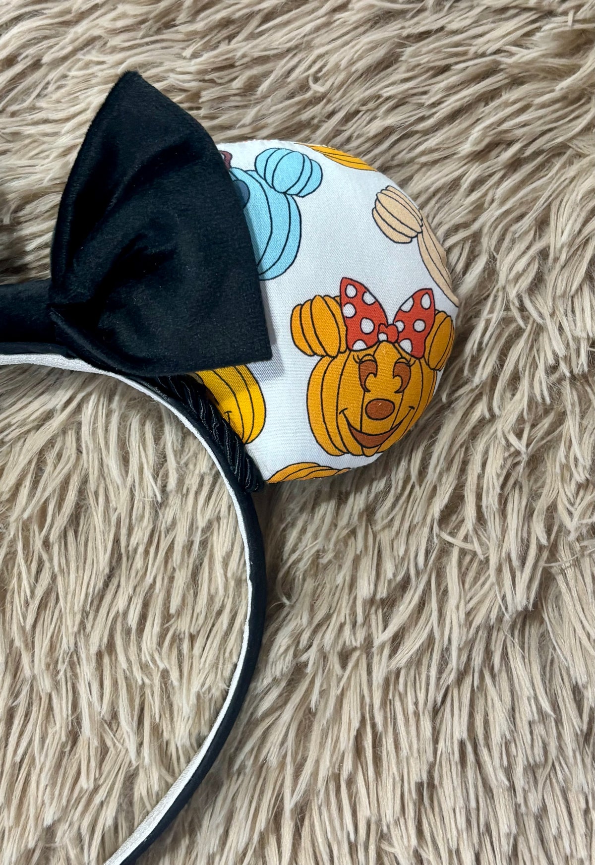 Pumpkin-themed ear headband featuring a light white background with orange, yellow, and blue pumpkins, a large sequined orange bow, and a black headband.