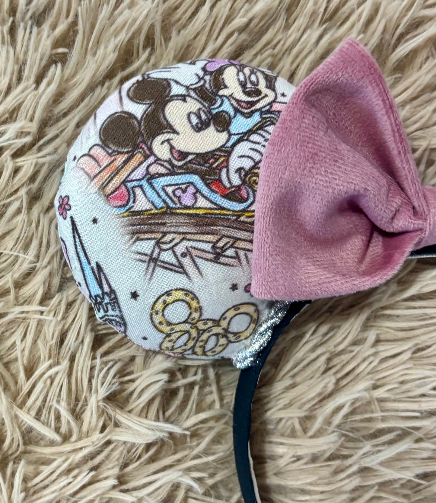 Close up of A pair of handmade ear headbands with a large dusty pink velvet bow, featuring printed park themed patterns on the ears and resting on a beige faux fur surface.