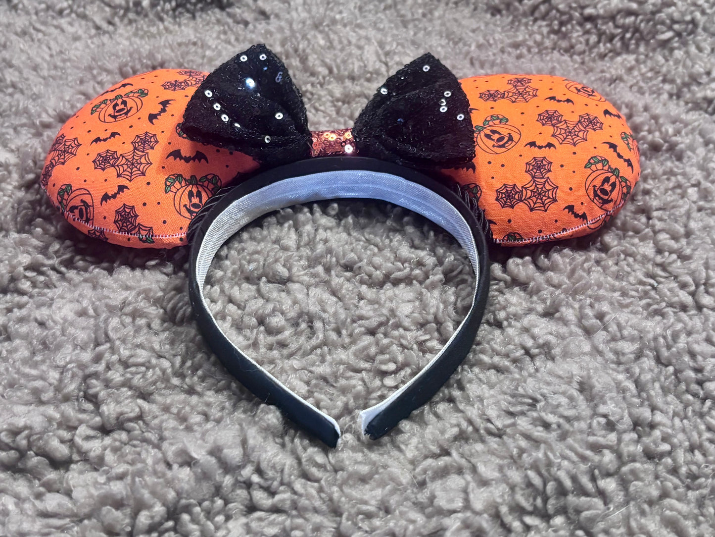 Halloween mouse ears with the fabric on both the front and back. It's orange with bats, mouse shaped pumpkins and spider webs. The headband is black universal sized and fits most. The set has a black with orange middle sequin bow. Item is shown on a brown furry background.
