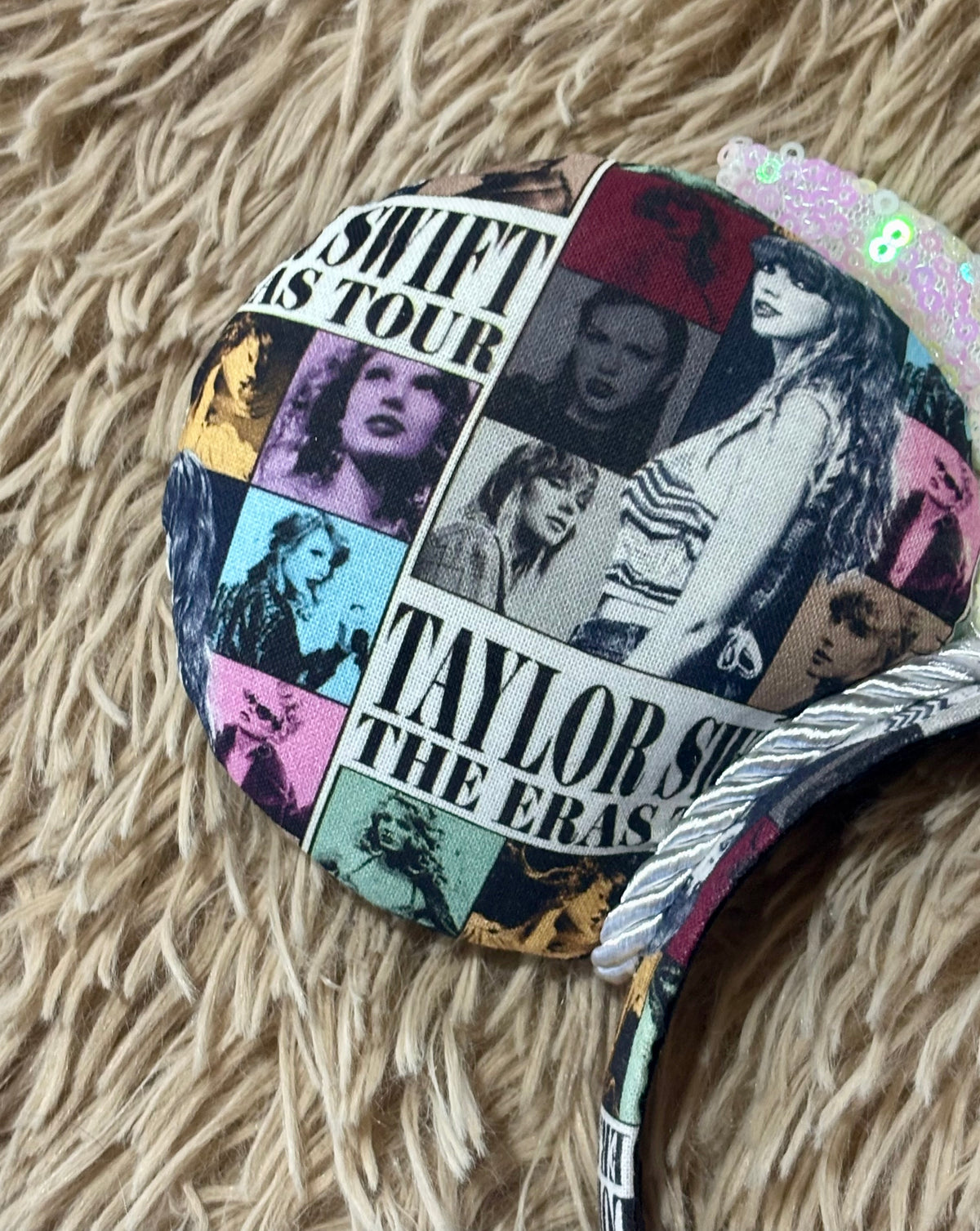 A close-up of a round fabric headband featuring a collage print of TS. The headband is placed on a beige faux fur surface.