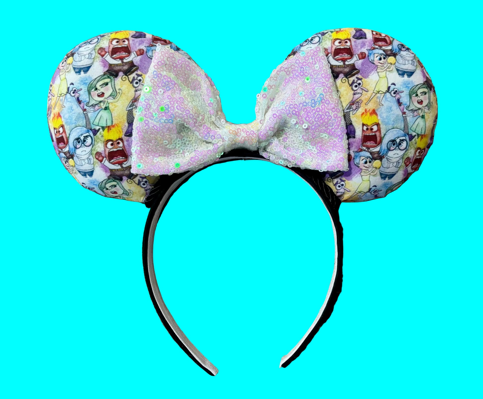 A pair of mouse ears headband featuring a colorful cartoon character print on the ears and a large iridescent glitter bow in the center, displayed on a beige shag rug.