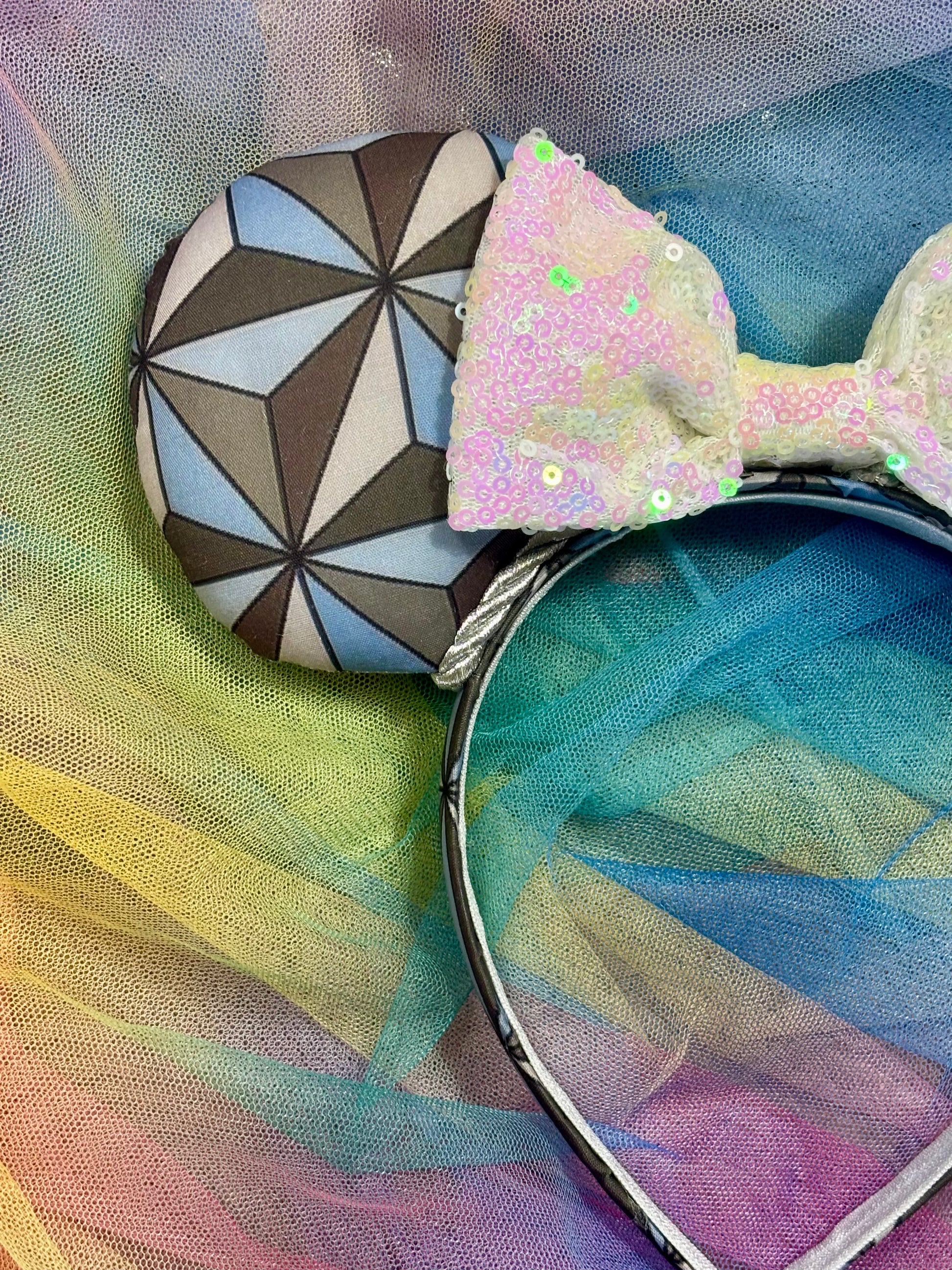 Mouse ear headband with a geometric design inspired by Spaceship Earth ride and a white iridescent bow on a colorful background. Shown from a close view of front mouse ear.