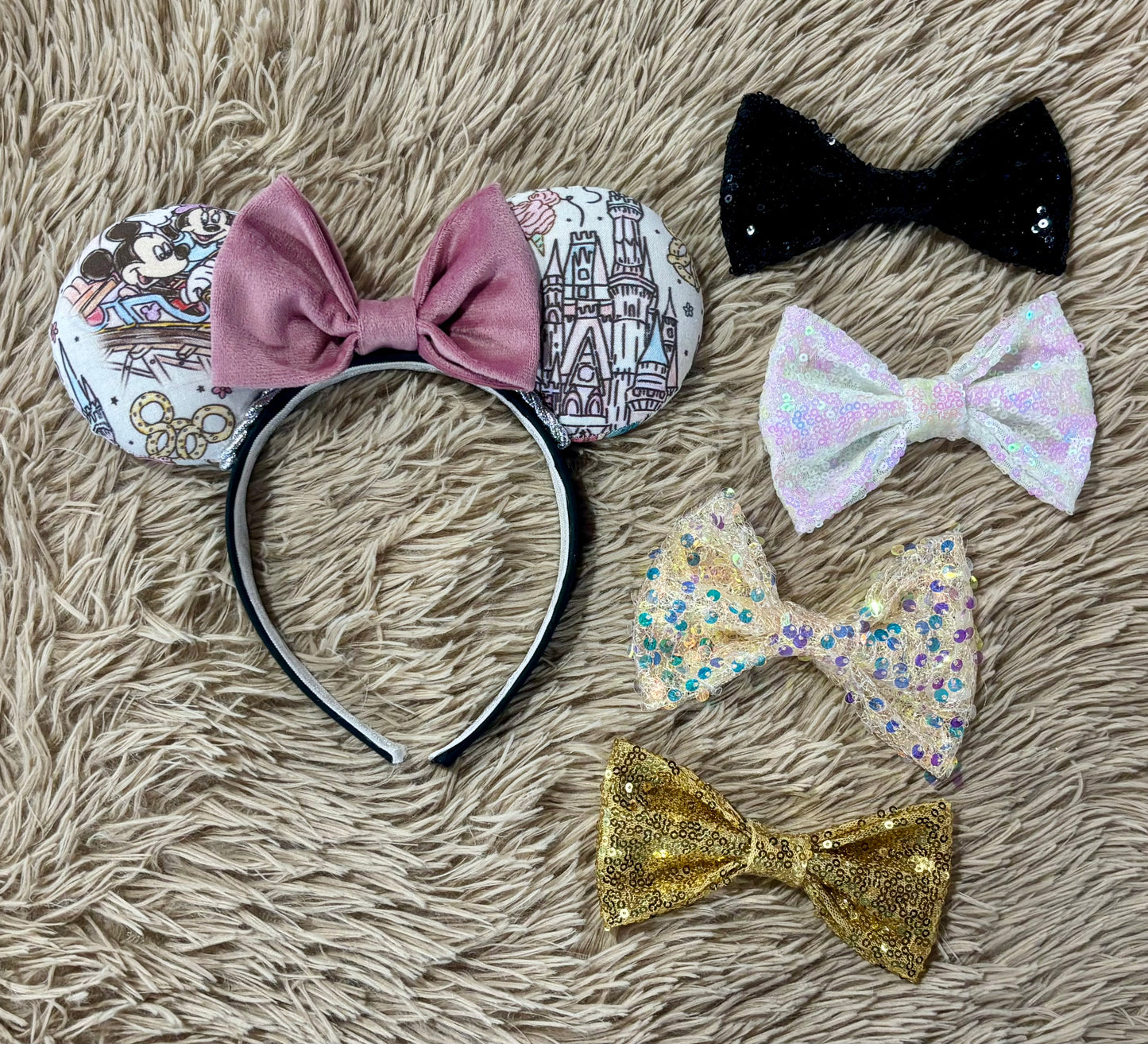 A pair of handmade ear headbands with a large dusty pink velvet bow, featuring printed park themed patterns on the ears and resting on a beige faux fur surface.