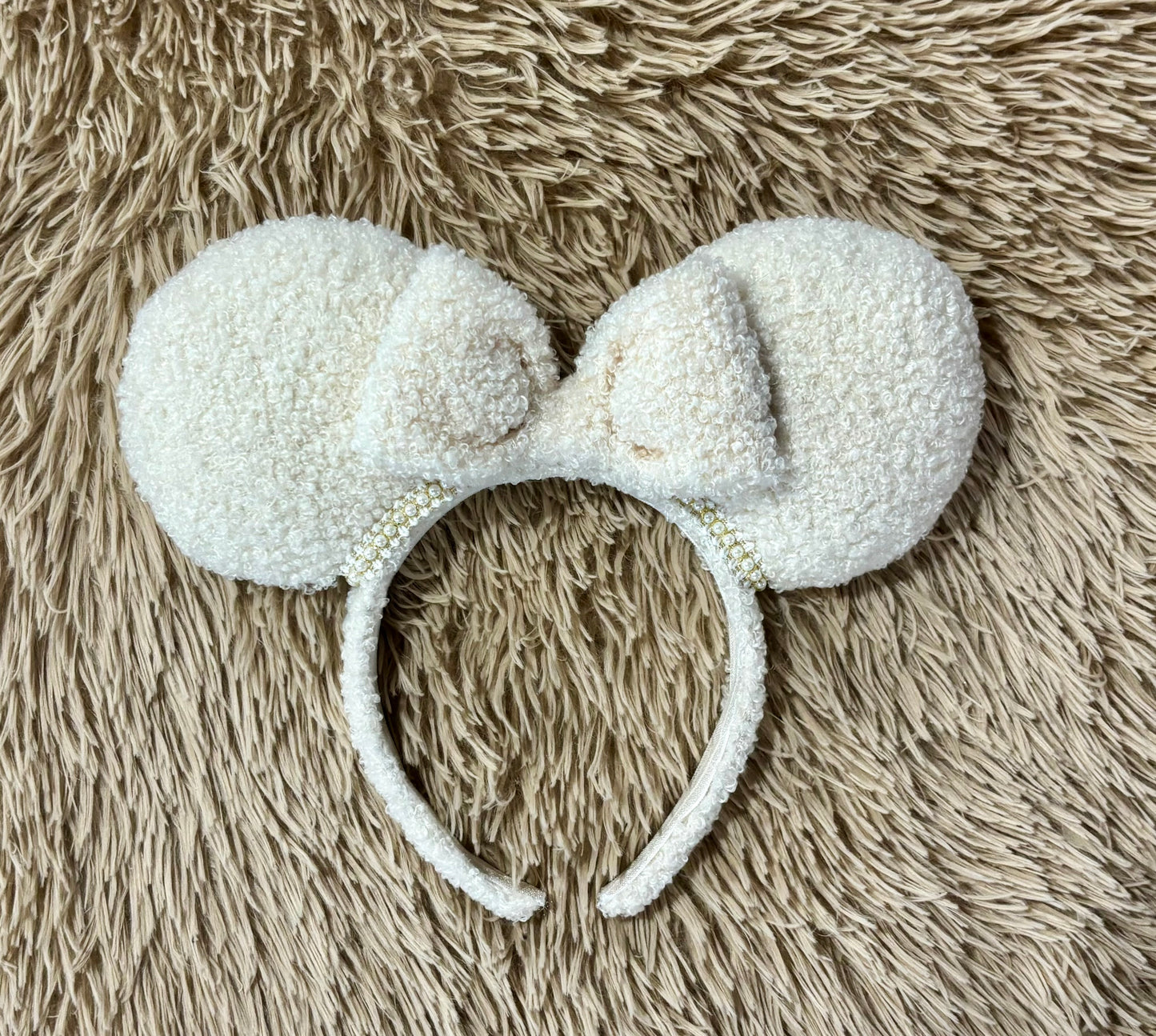 A cream-colored fuzzy ear headband with a bow, with pearl trim along the inner ears. Set is shown laid flat on a textured beige faux fur background.