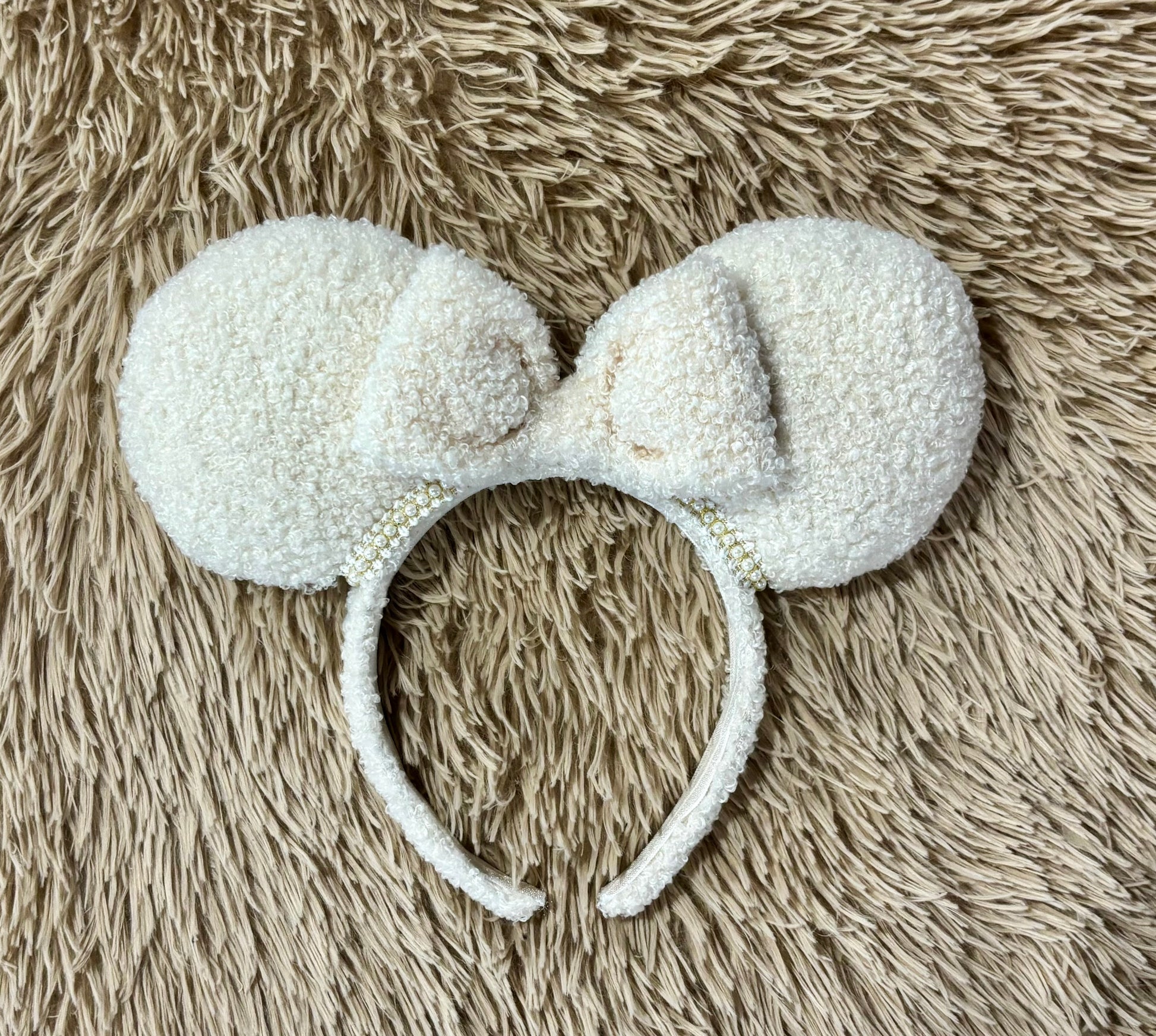 A cream-colored fuzzy ear headband with a bow, with pearl trim along the inner ears. Set is shown laid flat on a textured beige faux fur background.