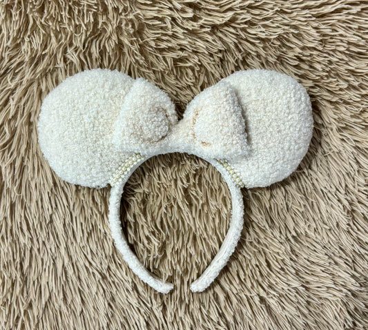 A cream-colored fuzzy ear headband with a bow, with pearl trim along the inner ears. Set is shown laid flat on a textured beige faux fur background.