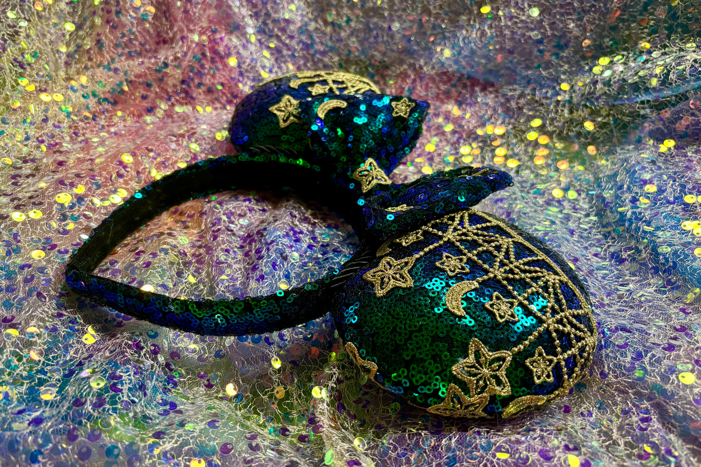 Mouse Ear headband that is a Handmade Iridescent blue and green sequins with gold sun, moon and stars attached to them. bow with star and moon patterns on a glittery background.