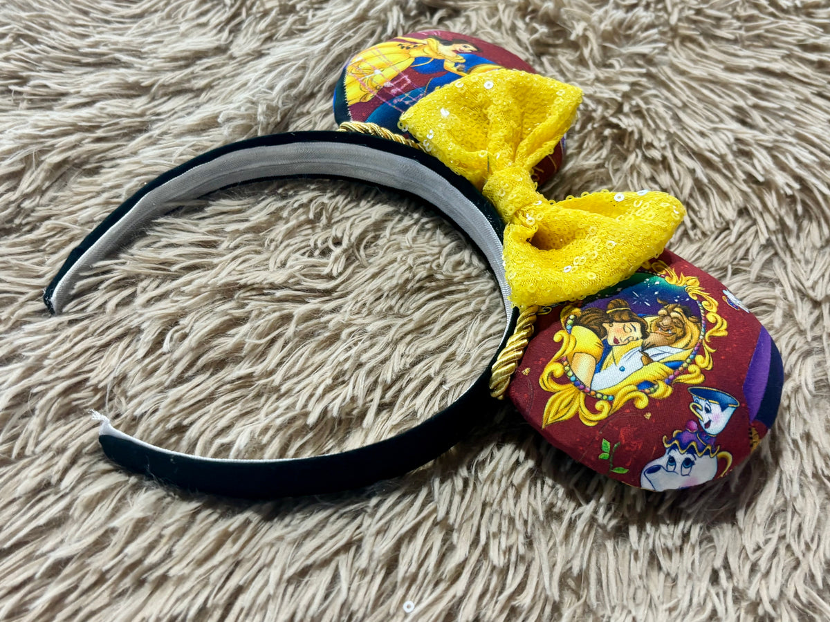 A headband featuring a large yellow glitter bow and a round printed charm with Belle and the beast show on a beige shag rug.