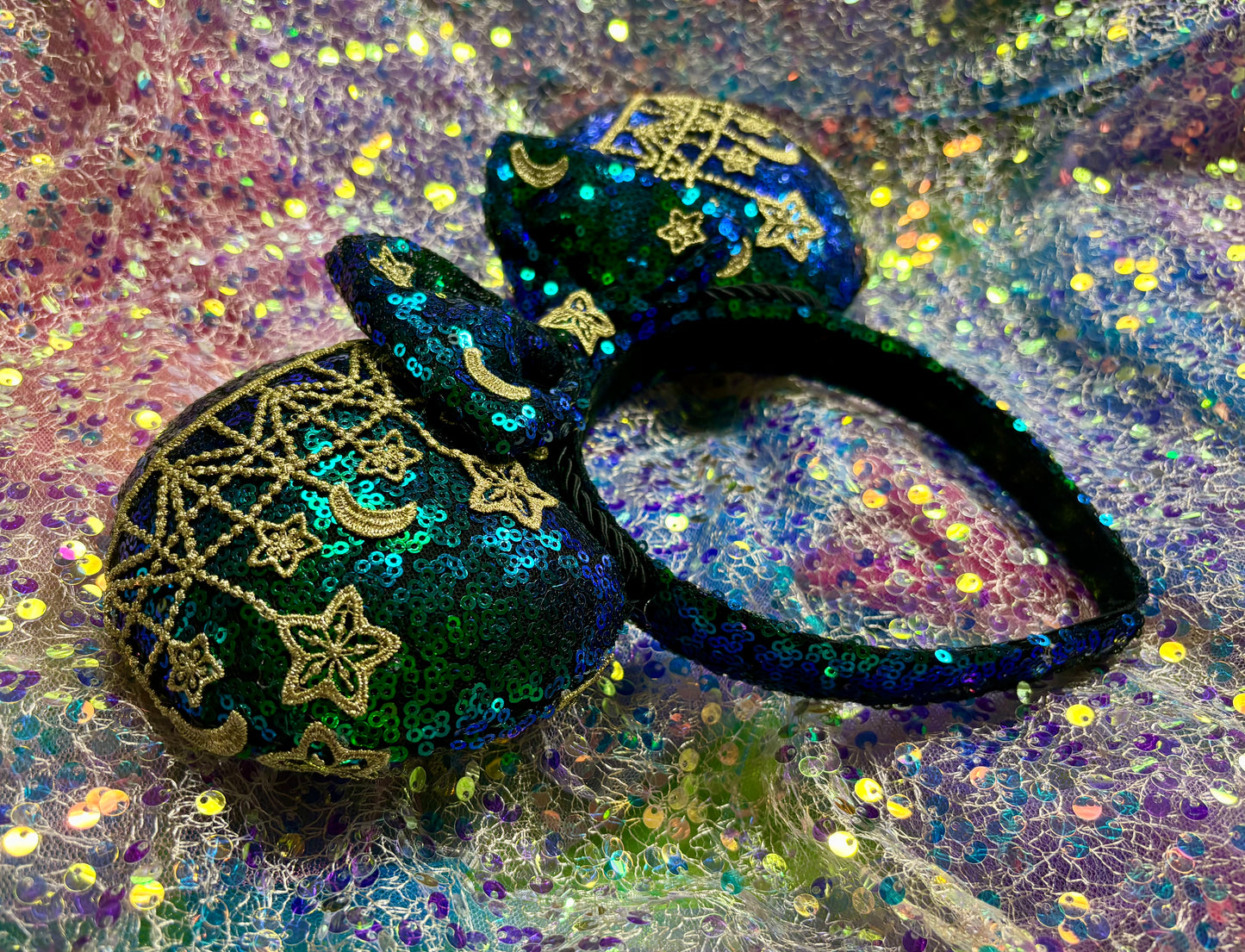 Mouse Ear headband that is a Handmade Iridescent blue and green sequins with gold sun, moon and stars attached to them. bow with star and moon patterns on a glittery background.