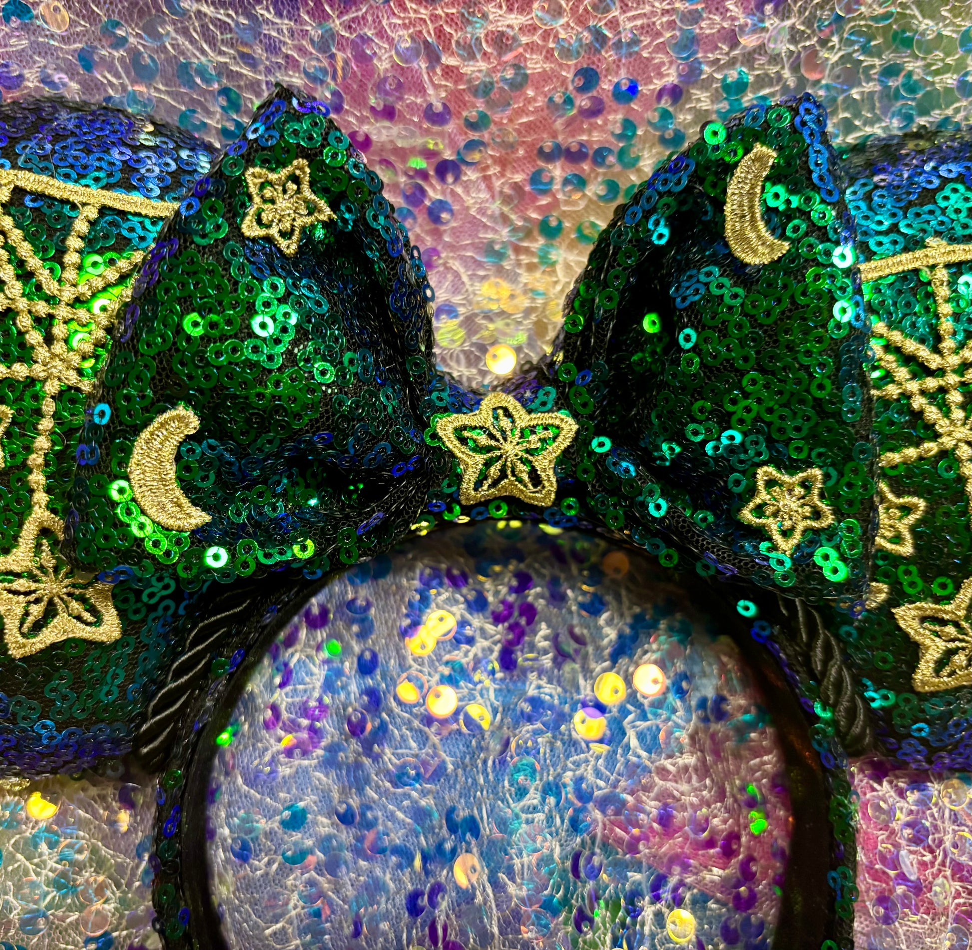 Mouse Ear headband that is a Handmade Iridescent blue and green sequins with gold sun, moon and stars attached to them. bow with star and moon patterns on a glittery background.