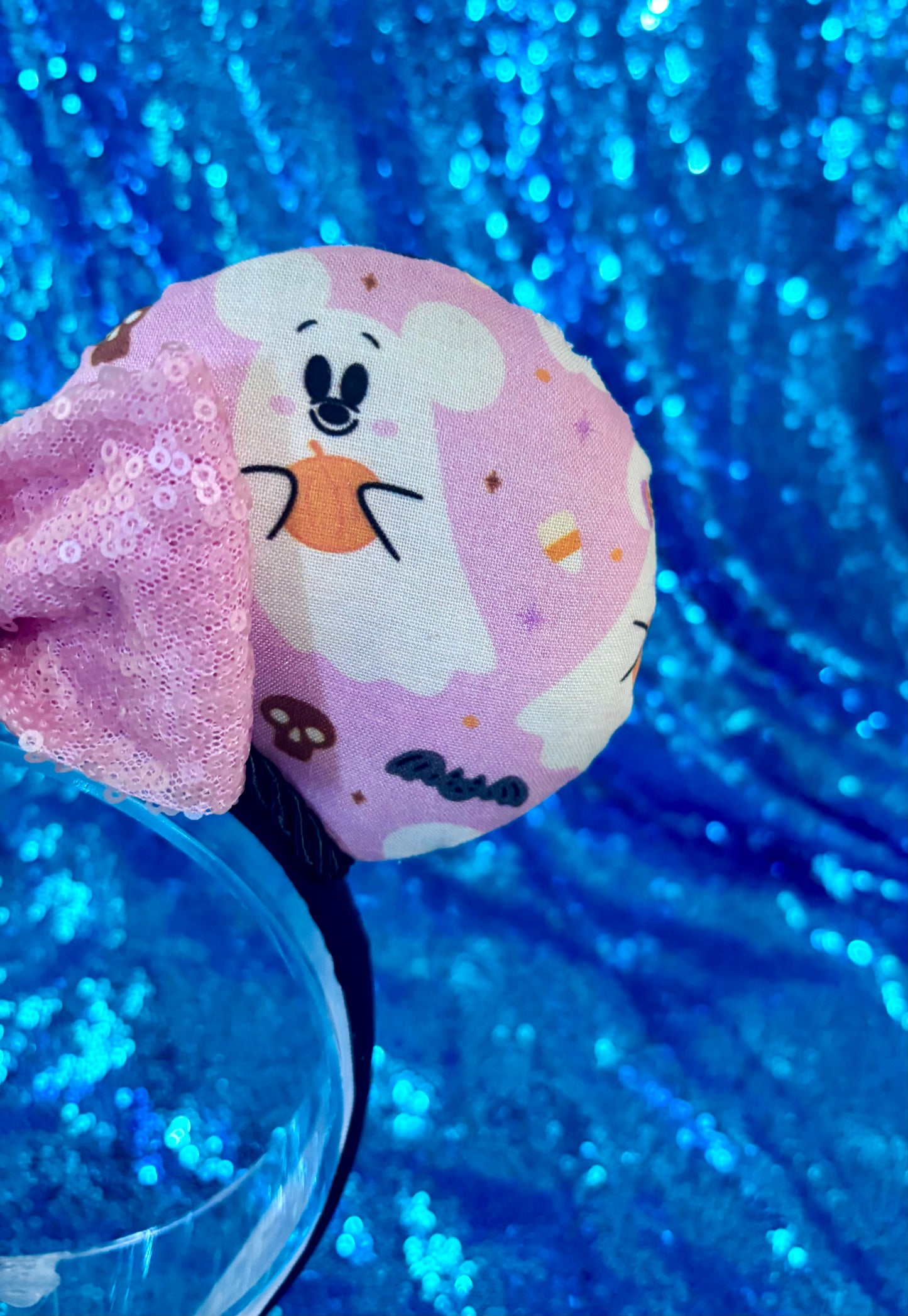 Cartoon character ghosts on a mouse ear headband with pink sequin bow shown on a blue glittery background.