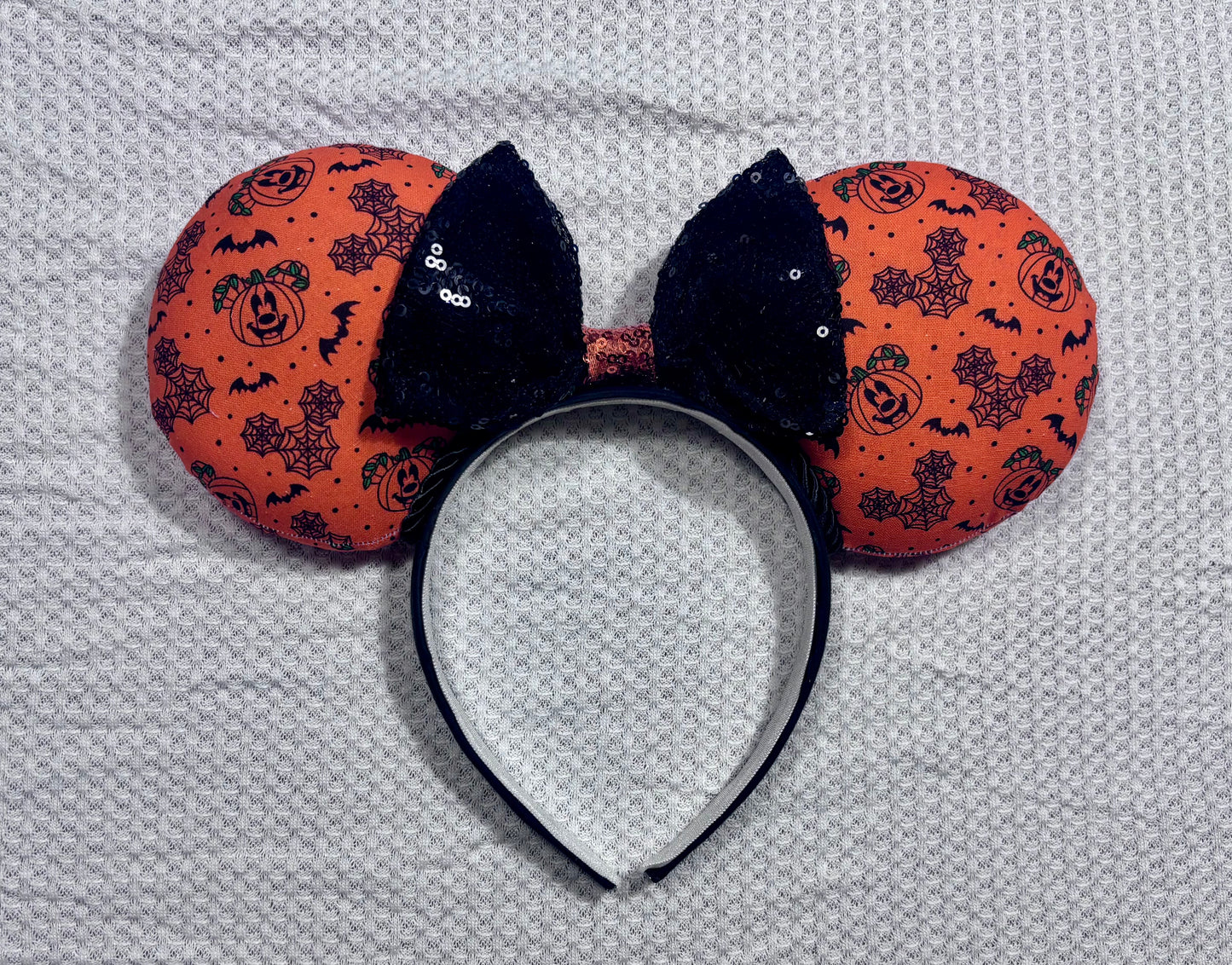 Halloween mouse ears with the fabric on both the front and back. It's orange with bats, mouse shaped pumpkins and spider webs. The headband is black universal sized and fits most. The set has a black with orange middle sequin bow. Item is shown on a white background.