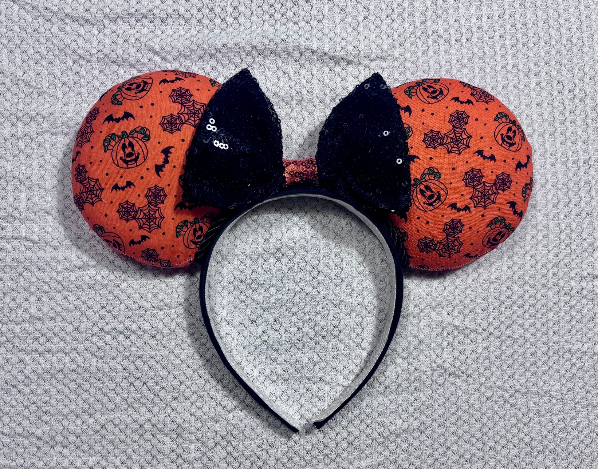 Halloween mouse ears with the fabric on both the front and back. It's orange with bats, mouse shaped pumpkins and spider webs. The headband is black universal sized and fits most. The set has a black with orange middle sequin bow. Item is shown on a white background.
