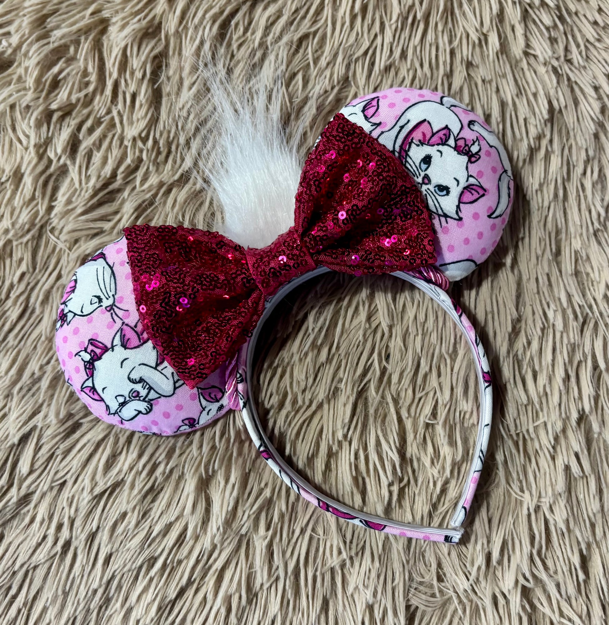 A pair of Marie inspired ears featuring pink fabric with white cartoon cats and pink polka dots, a large hot pink sequined bow, and a white faux fur accent, displayed on a beige shag rug.