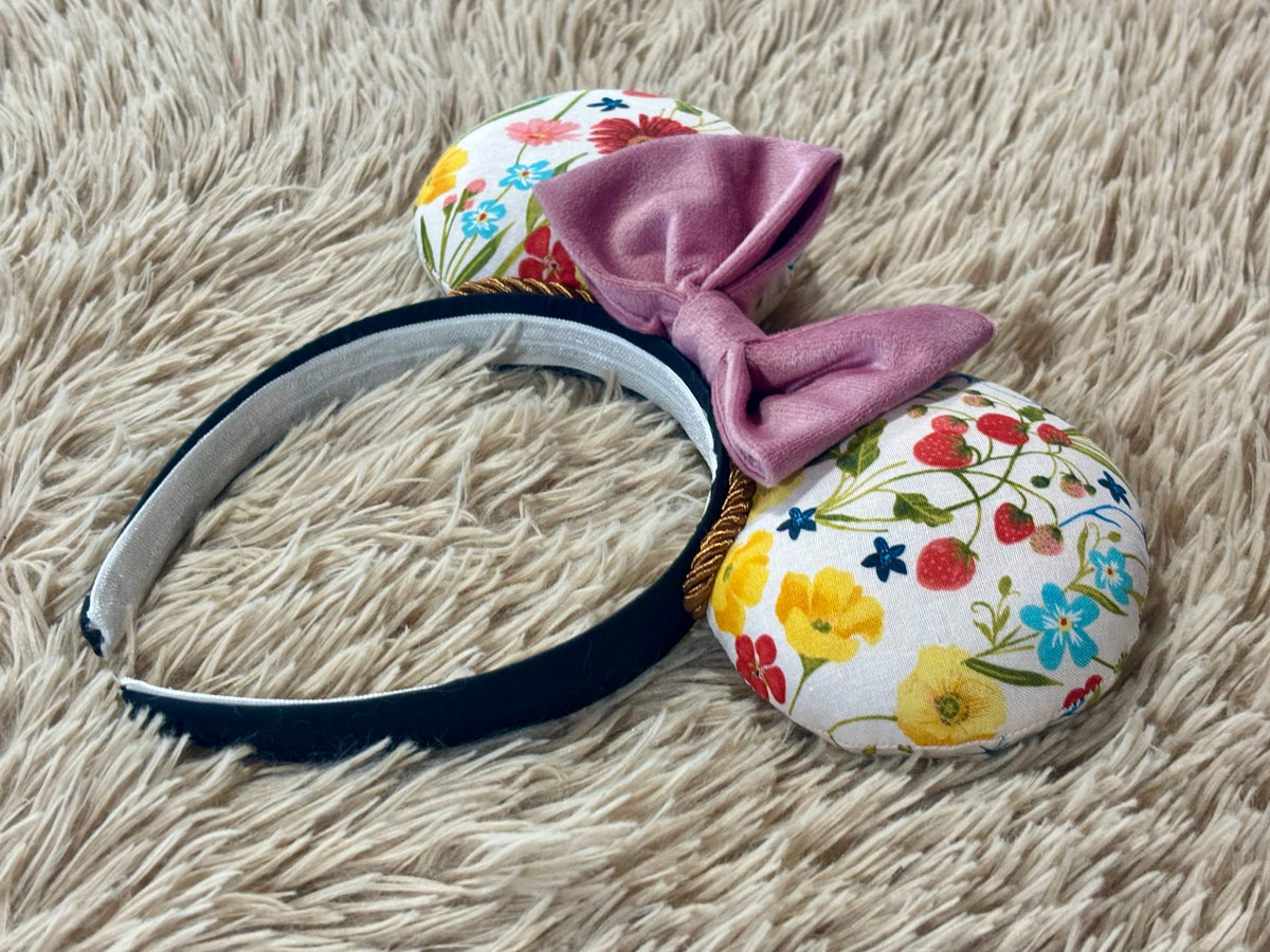 A handmade headband featuring two round, fabric-covered ears with a colorful floral print on a white background, a dusty pink velvet bow, and a black band.