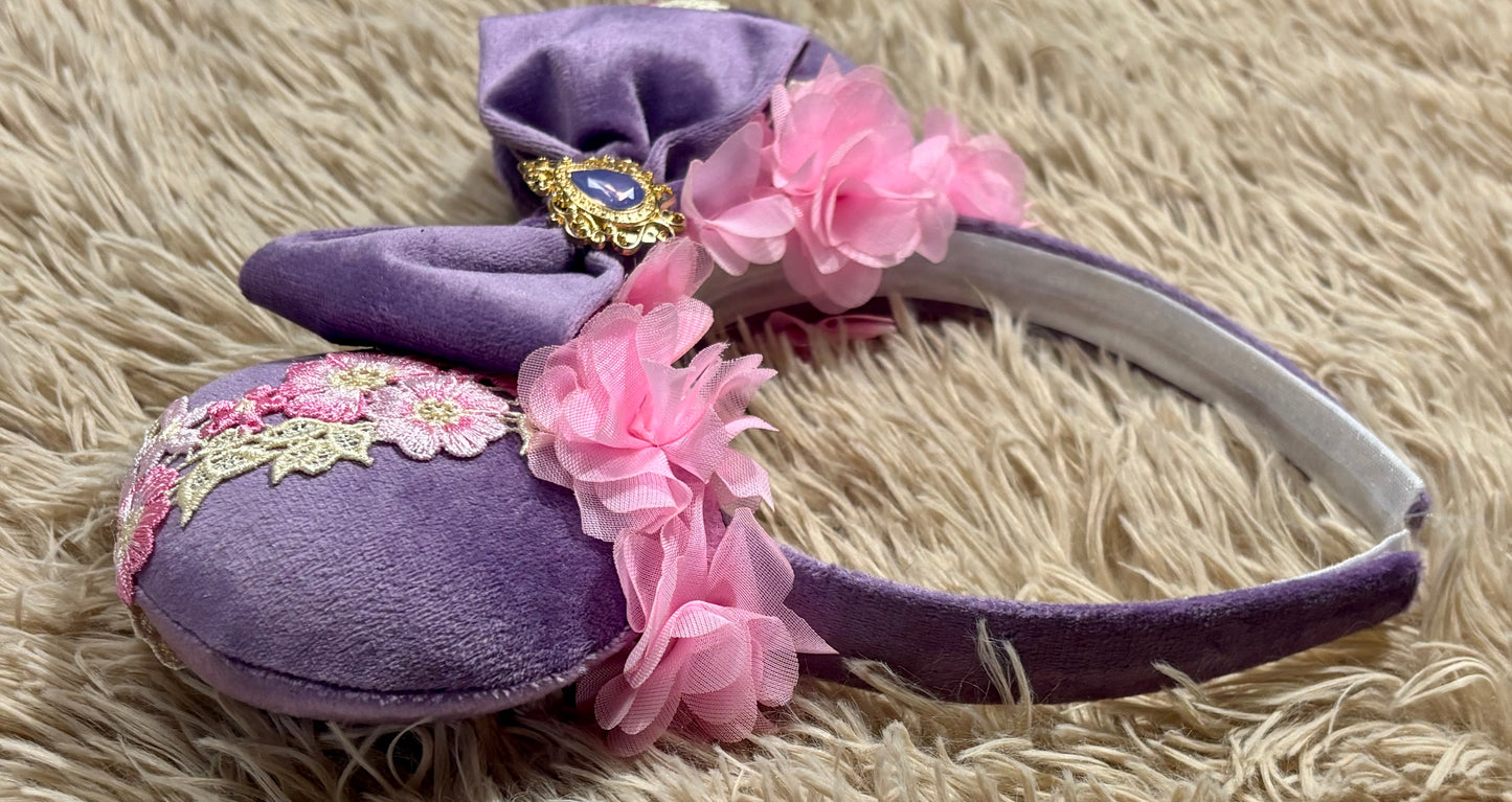 Purple and pink fabric mouse ears headband with a large velvet bow, floral lace appliques, and a purple gem accent.