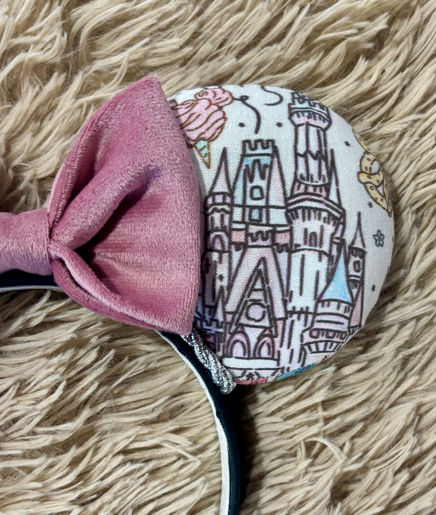 A close-up of a fabric-covered headband featuring a large dusty pink bow and a round white section printed with a castle illustration and pastel accents, resting on a beige shag rug.