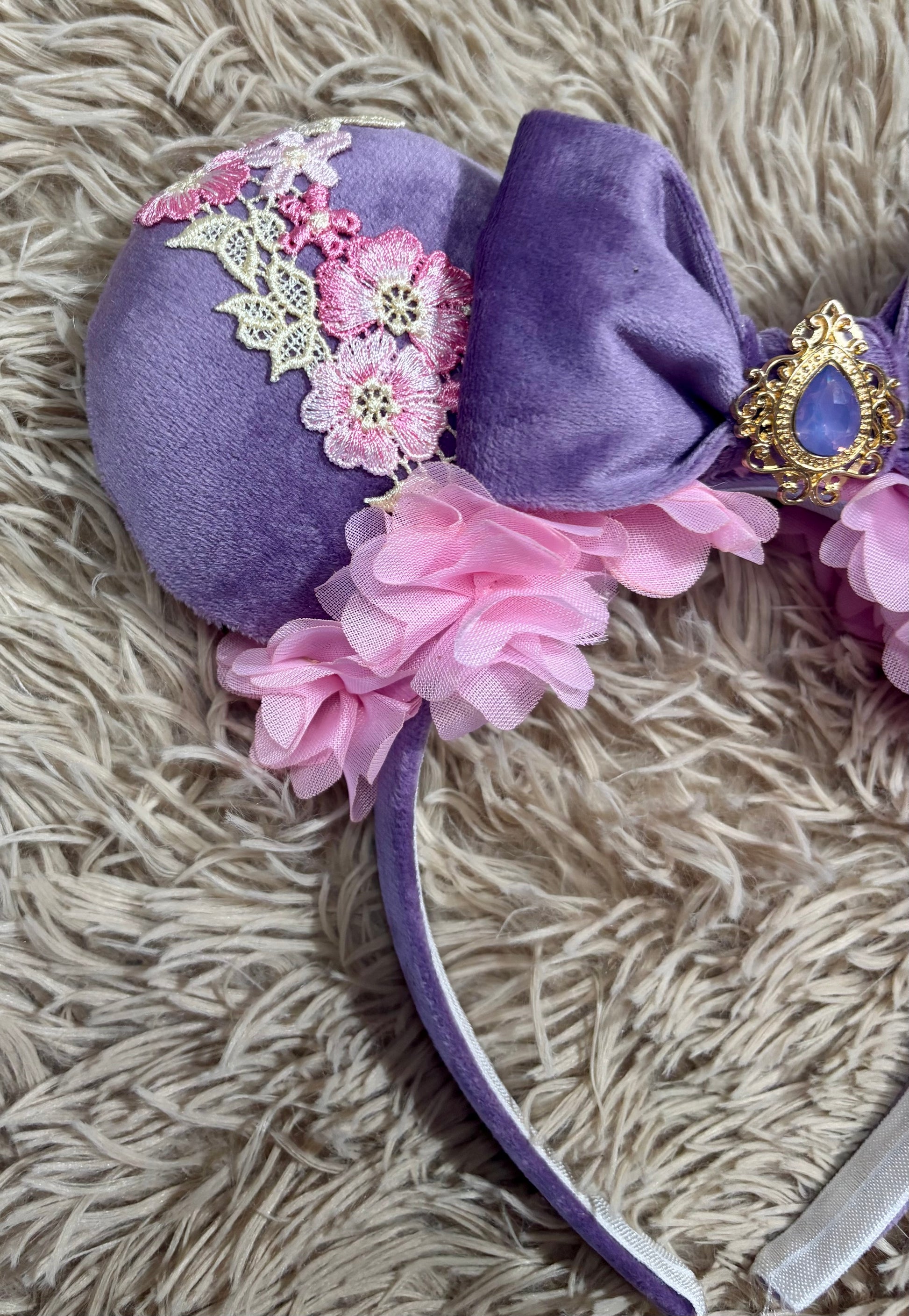 A purple velvet headband featuring a large bow, pink fabric flowers, white lace applique, and a gold ornate jewel accent, laid flat on a beige shag rug.