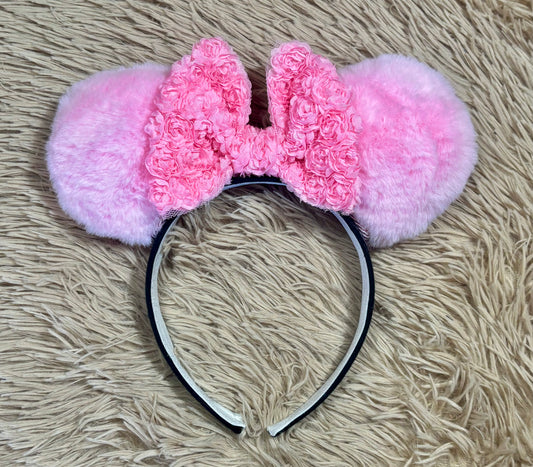 Pink fuzzy ears headband with a textured pink bow and a black headband, displayed on a beige shag rug.