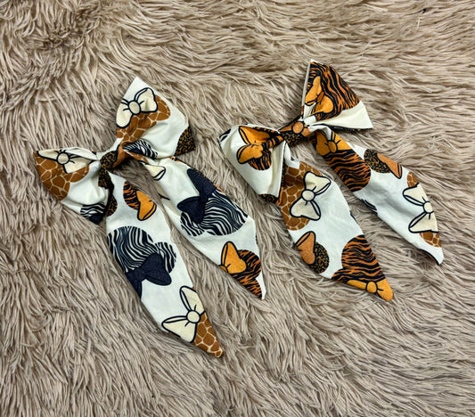 Two fabric hair bows with animal and nature-inspired prints, displayed on a shaggy beige faux fur background. The left bow features giraffe, zebra, and butterfly motifs on a white base, while the right bow features tiger, giraffe, and butterfly patterns.