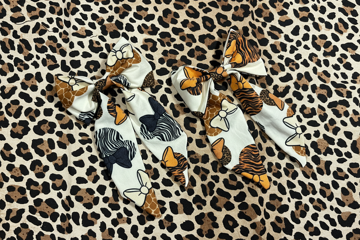Two fabric hair bows with animal and nature-inspired prints, displayed on a leopard print background. The left bow features giraffe, zebra, and tiger motifs on a white base, while the right bow features tiger, giraffe, and cheetah patterns.