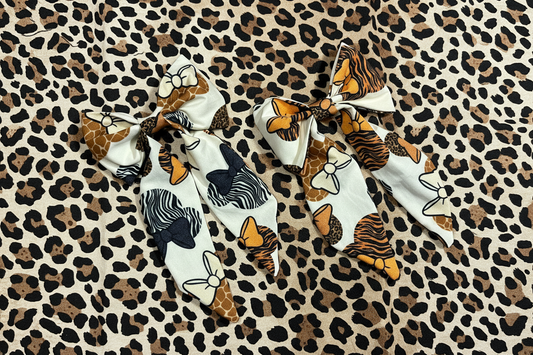 Two fabric hair bows with animal and nature-inspired prints, displayed on a leopard print background. The left bow features giraffe, zebra, and tiger motifs on a white base, while the right bow features tiger, giraffe, and cheetah patterns.