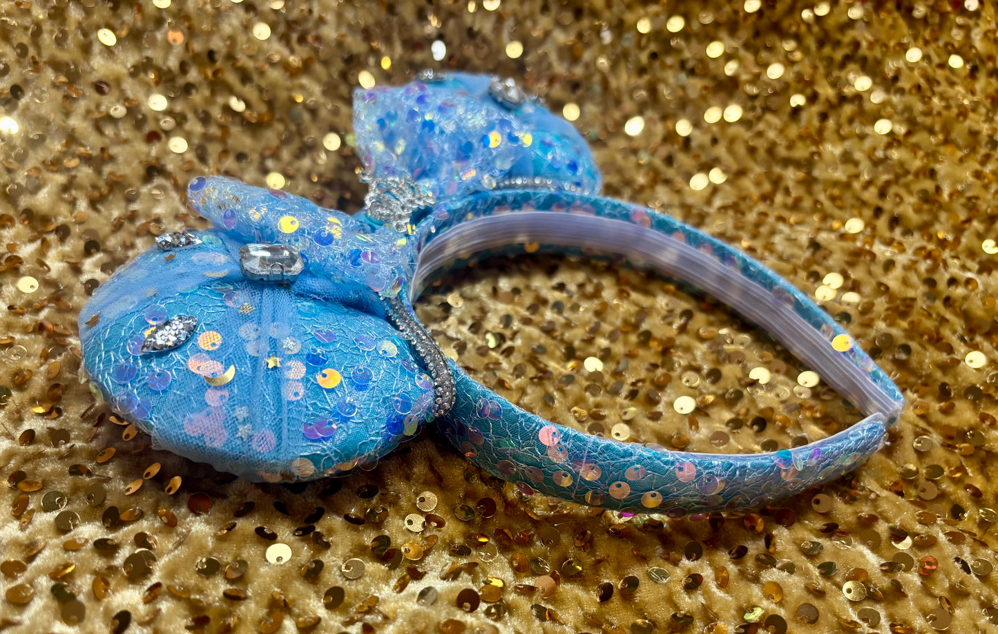 Handmade and ready to ship blue sequin mouse ears headband inspired by Cinderella with tulle and silver gemstones with a crown charm middle and rhinestone trim along inner ears. Close up of side view of headband.