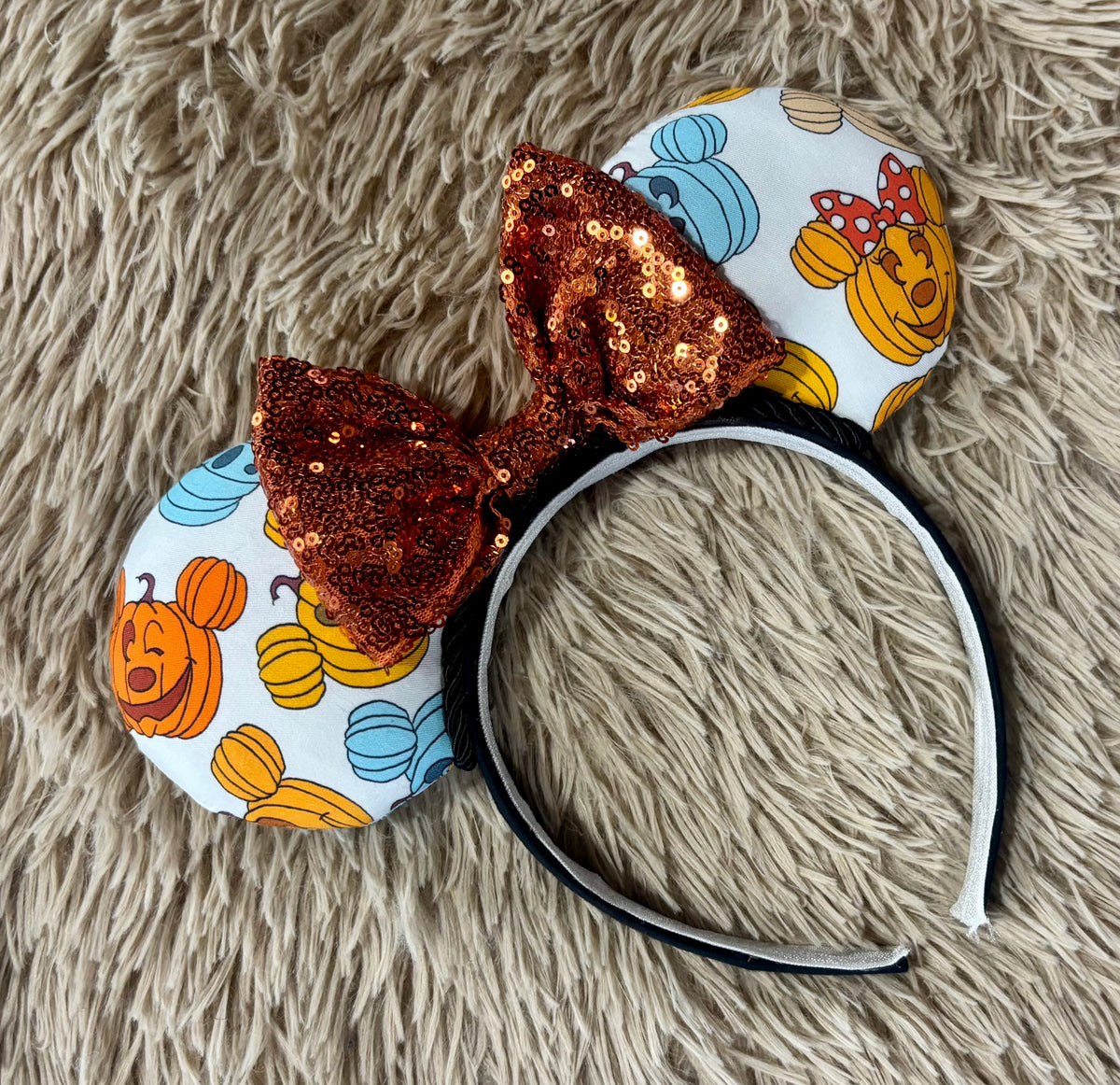 A pair of mouse ears headband featuring a white fabric print with orange pumpkins, blue acorns, and a large sequined brown bow, displayed on a beige shag rug.