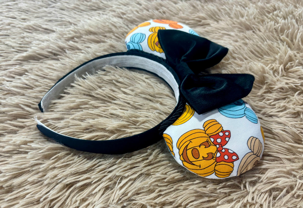 A headband with ears covered in a colorful pumpkin cartoon print and a large black bow, resting on a beige shag rug.