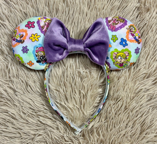 A pair of ear headband accessories with a large purple velvet bow in the center and multicolored Lizzie style printed ears, displayed on a beige faux-fur surface.